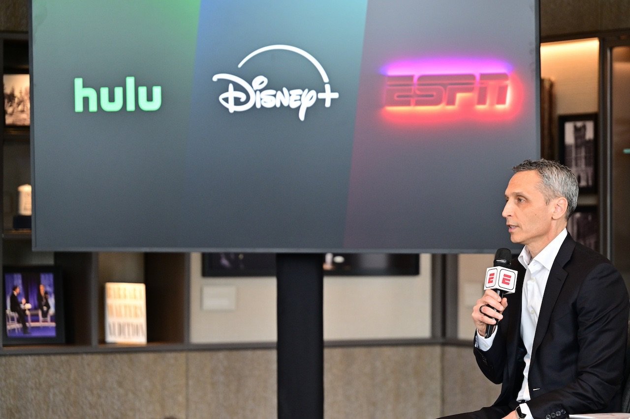 Ketua ESPN berbicara dengan mikrofon di depan TV menunjukkan saluran streaming yang berbeda